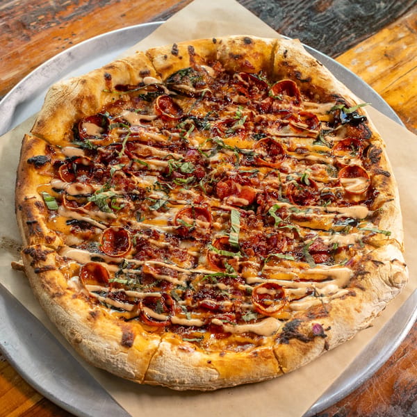 Delicious pepperoni and bacon pizza with basil and creamy sauce drizzle on a rustic wooden table.