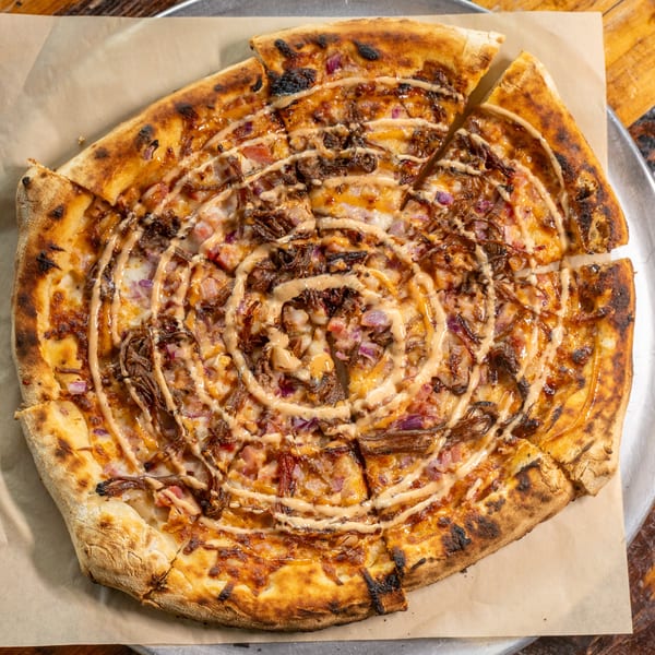 Wood-fired BBQ pulled pork pizza with red onions and sauce drizzle on a metal tray.
