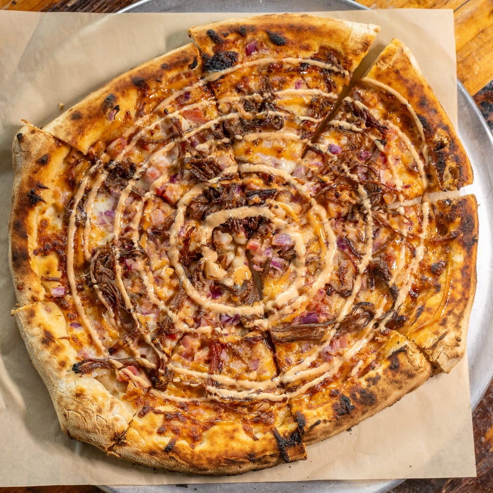 Wood-fired BBQ pulled pork pizza with red onions and sauce drizzle on a metal tray.