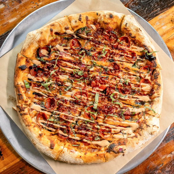 Whole pizza with pepperoni, bacon, basil, and sauce drizzle on a metal tray and wooden table.