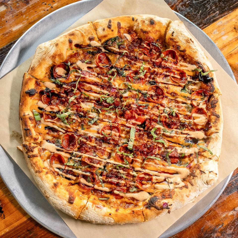 Whole pizza with pepperoni, bacon, basil, and sauce drizzle on a metal tray and wooden table.