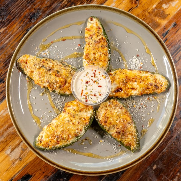 Five cheesy baked jalapeño halves arranged in a star around a dipping sauce cup on a grey plate.